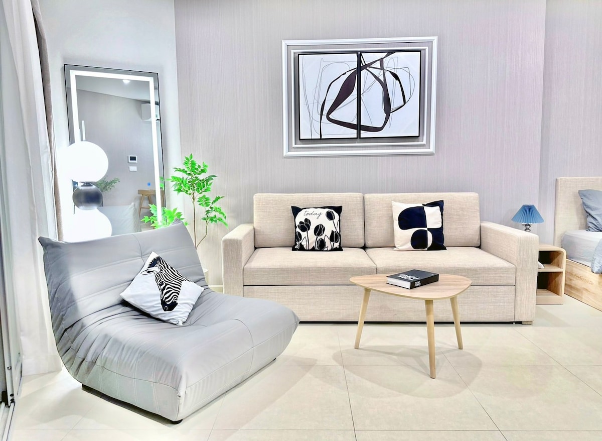 A light-filled living area features a modern sofa with decorative pillows and a sleek armchair. A round coffee table sits centrally, while a piece of abstract wall art adds visual interest. A large mirror reflects the space, enhancing its openness.