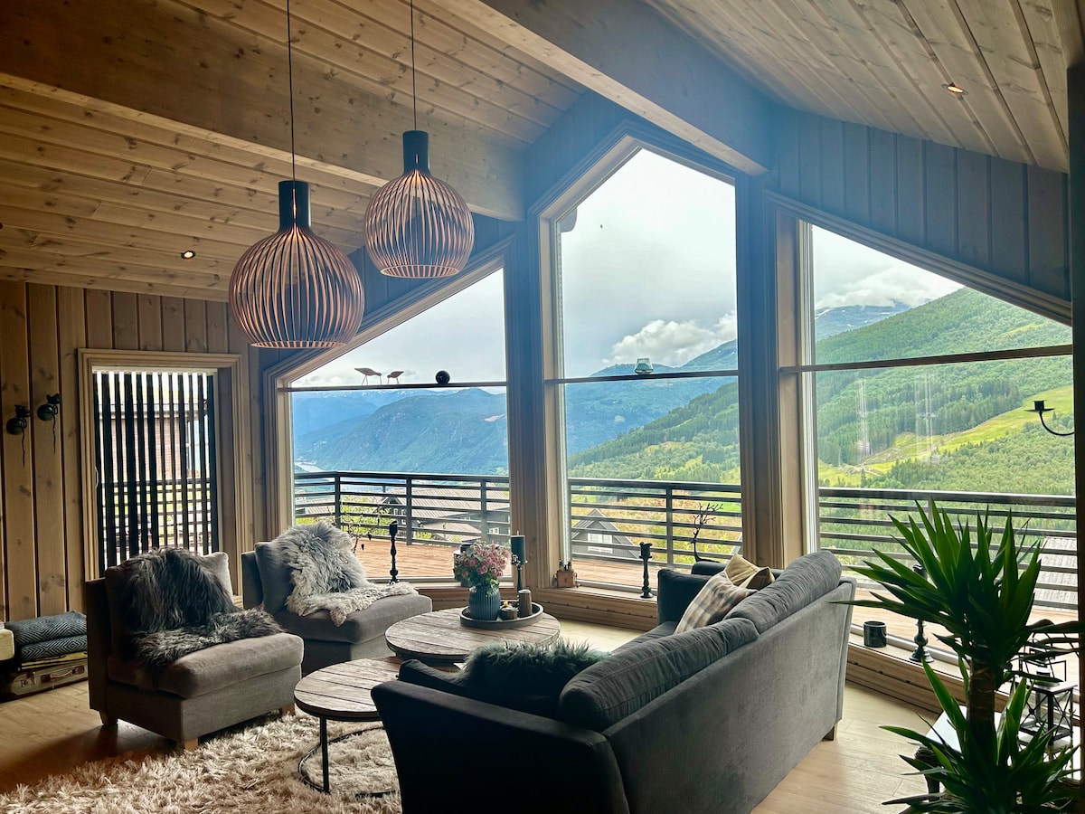 A spacious living area features geometric windows that frame scenic mountain views. Comfortable seating is arranged around a central coffee table, with soft textiles adding texture. Natural light floods the space, enhancing the warm wooden interior.