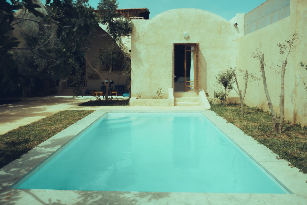 An inviting outdoor area features a crystal-clear swimming pool surrounded by manicured grass. A pathway leads to an entrance with a large door, while trees provide subtle shade in the background, creating a serene environment for relaxation.