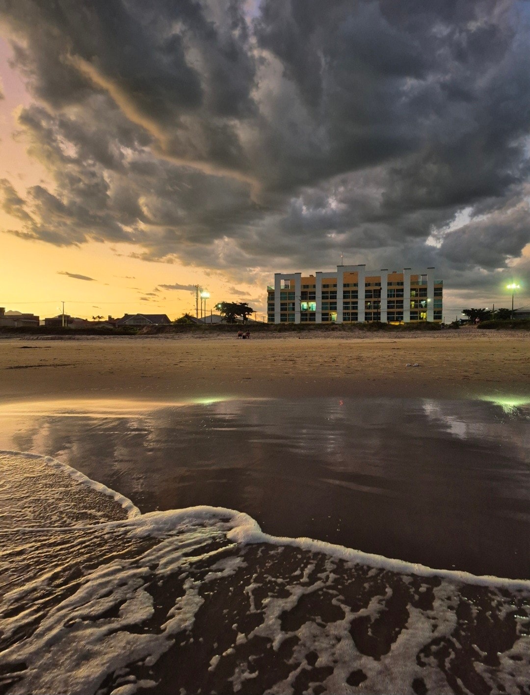 The building is positioned near the shoreline, with an evening sky illuminated by a warm glow. Gentle waves reach the sandy beach, reflecting the colors of the sunset. Cloud formations add depth to the scene, enhancing the coastal atmosphere.
