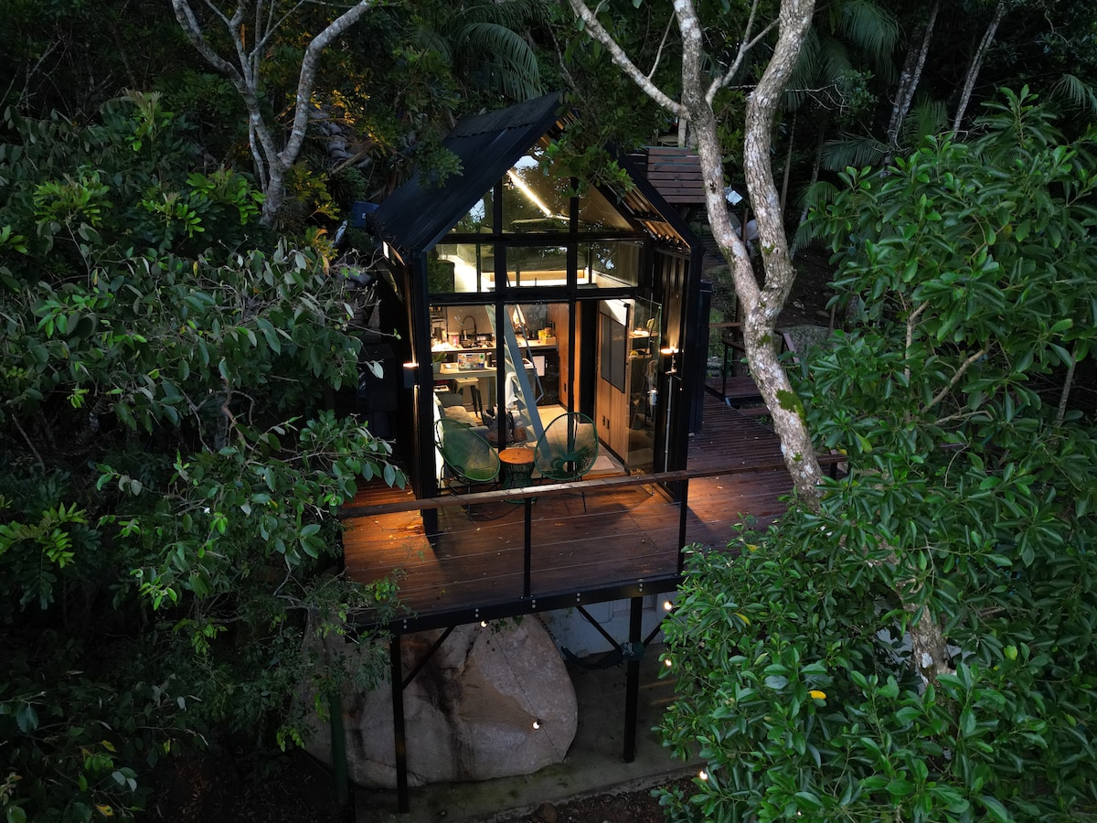 A tiny house is nestled among lush greenery, featuring a wooden deck and large glass windows. The interior is illuminated, revealing a cozy living area with a dining table and chairs, seamlessly blending with the surrounding nature.