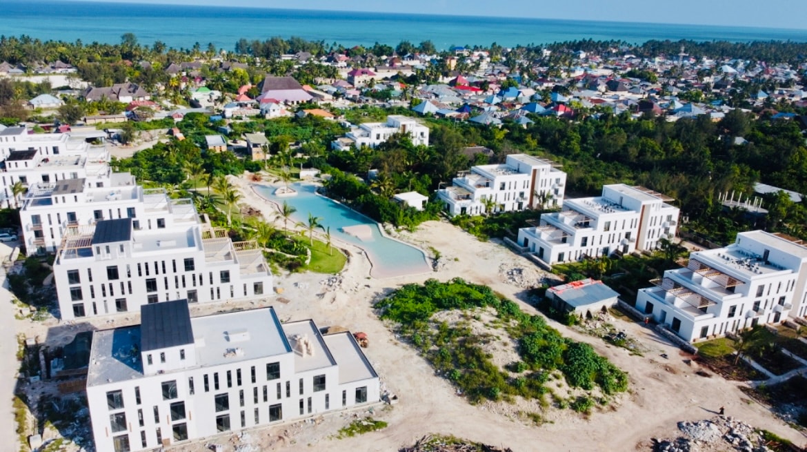 An aerial view reveals a serene landscape featuring a cluster of modern buildings within a gated community. A winding pool is visible amidst lush greenery, with the Indian Ocean in the background. The area shows an evolving residential environment with ongoing construction.