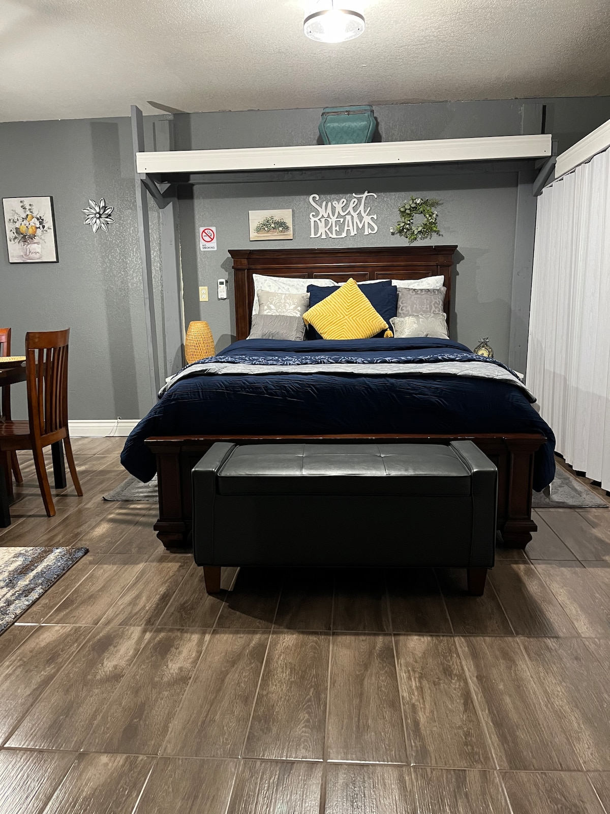The bedroom features a wooden bed with navy bedding and decorative pillows. A padded ottoman is positioned at the foot of the bed. A dining area with wooden chairs is visible nearby, along with a wall decorated with a 'Sweet Dreams' sign. Natural light enters through sheer curtains.
