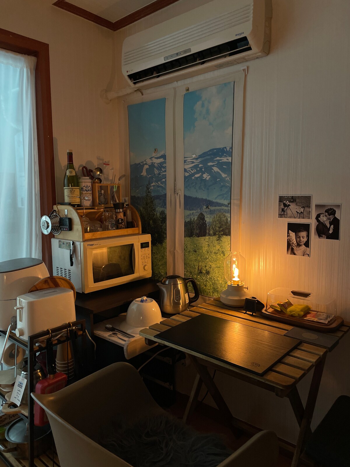 A compact kitchen area is presented, featuring a microwave, kettle, and a small table with various kitchen supplies. A window adorned with scenic wallpaper allows natural light, while a warm lamp provides illumination. The setting includes a few decorative photographs on the wall.