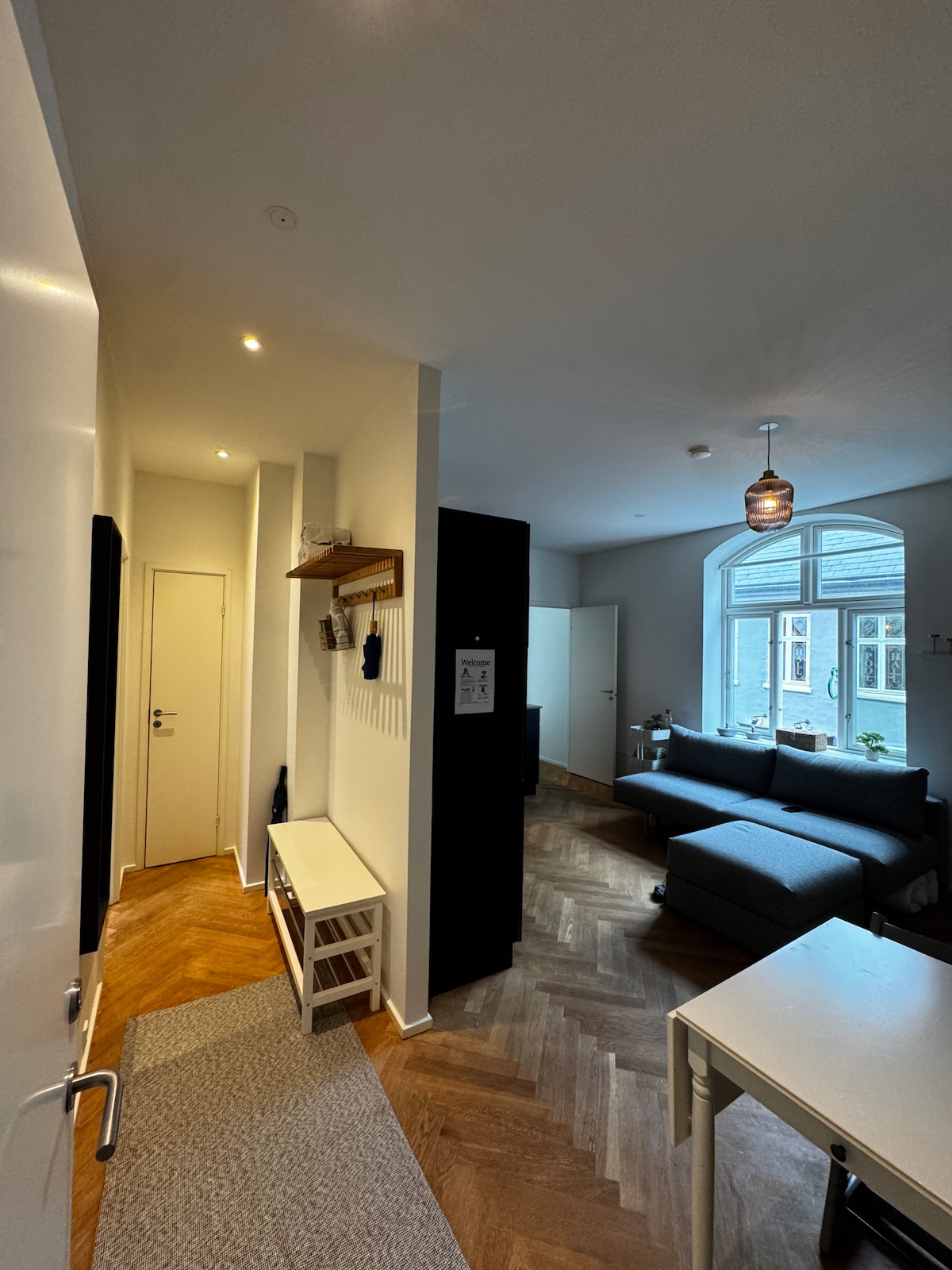 A well-lit entrance hallway features a minimalist design with a light wood bench and coat hooks. The adjoining living area is visible, showcasing a comfortable gray sofa and a dining table. Natural light filters through a large window, accentuating the warm wood flooring.