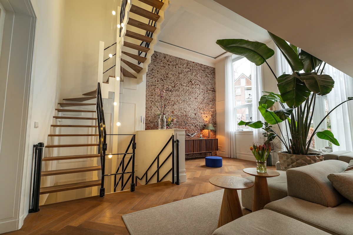 A bright and airy living space features an open staircase with wooden steps and a modern railing. Lush green plants are positioned near large windows, while warm wood flooring complements the brick accent wall. A comfortable sofa and coffee table add to the inviting ambiance.