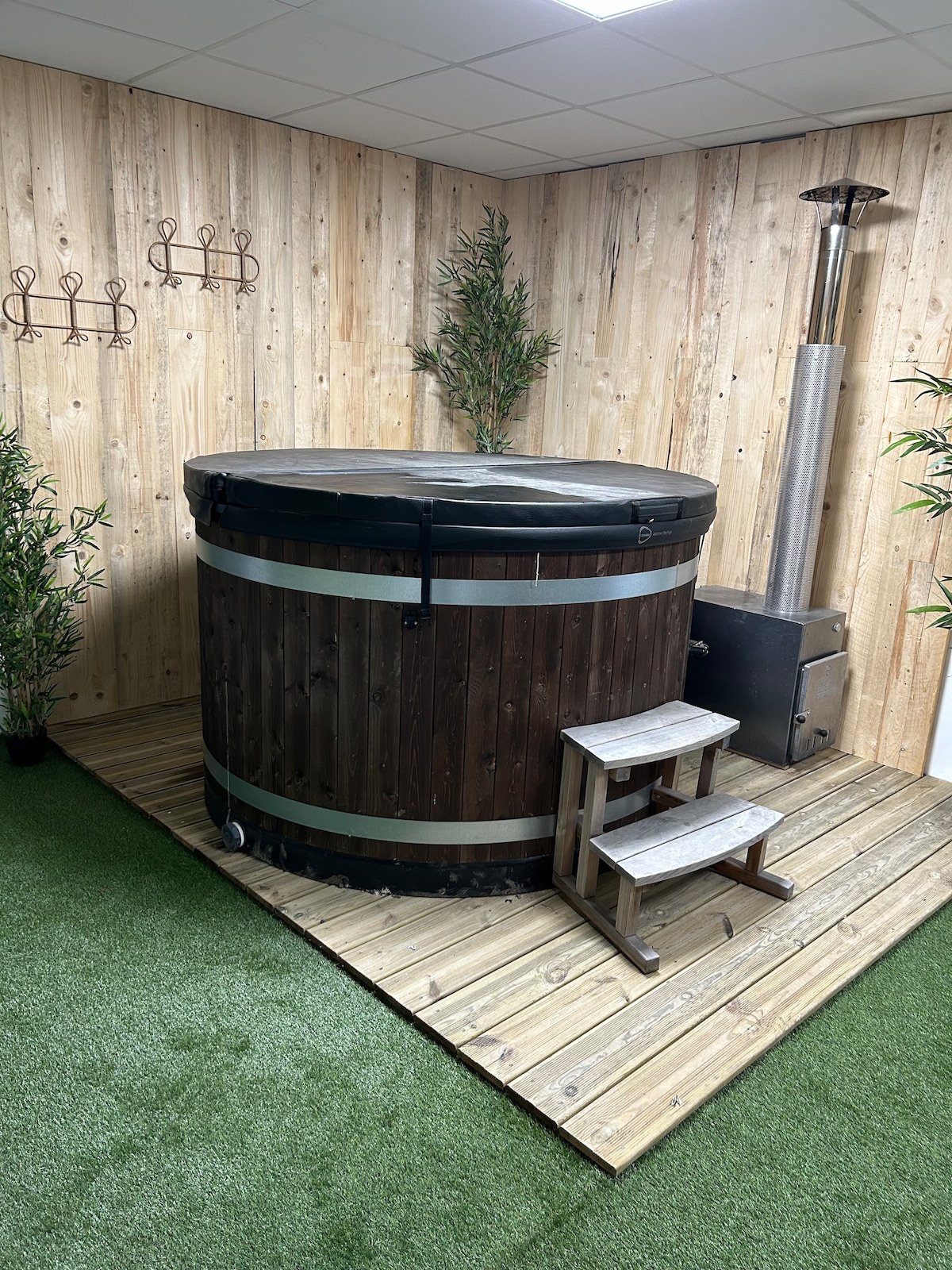 An indoor wooden hot tub with a black cover is positioned on a wooden deck. Two steps lead up to the tub, surrounded by artificial grass and minimal greenery. A metal stove stands beside the tub against a textured wooden wall.