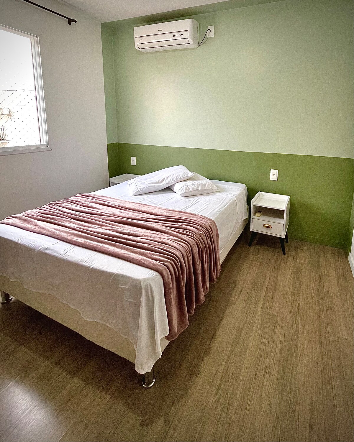A bright sleeping area features a spacious bed dressed in smooth white linens and accented by a soft pink throw. A small bedside table stands next to the bed, complementing the fresh green wall that enhances the overall calming atmosphere.