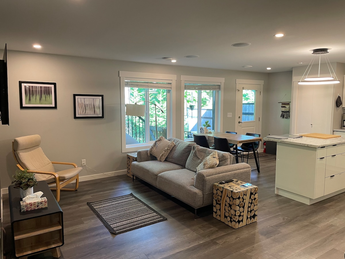 A modern living space is presented with large windows allowing natural light. A comfortable couch is accompanied by a chair and a small table made of logs. The dining area features a simple table and chairs, while artwork adorns the walls, complementing the sleek kitchen design.