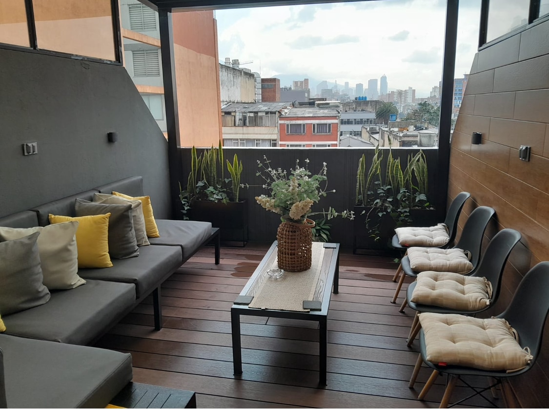 An outdoor terrace features a seating area with a grey couch adorned with yellow cushions. A wooden coffee table centers the space, topped with a decorative plant arrangement. Surrounding greenery enhances the tranquil atmosphere, while city views are visible in the background.