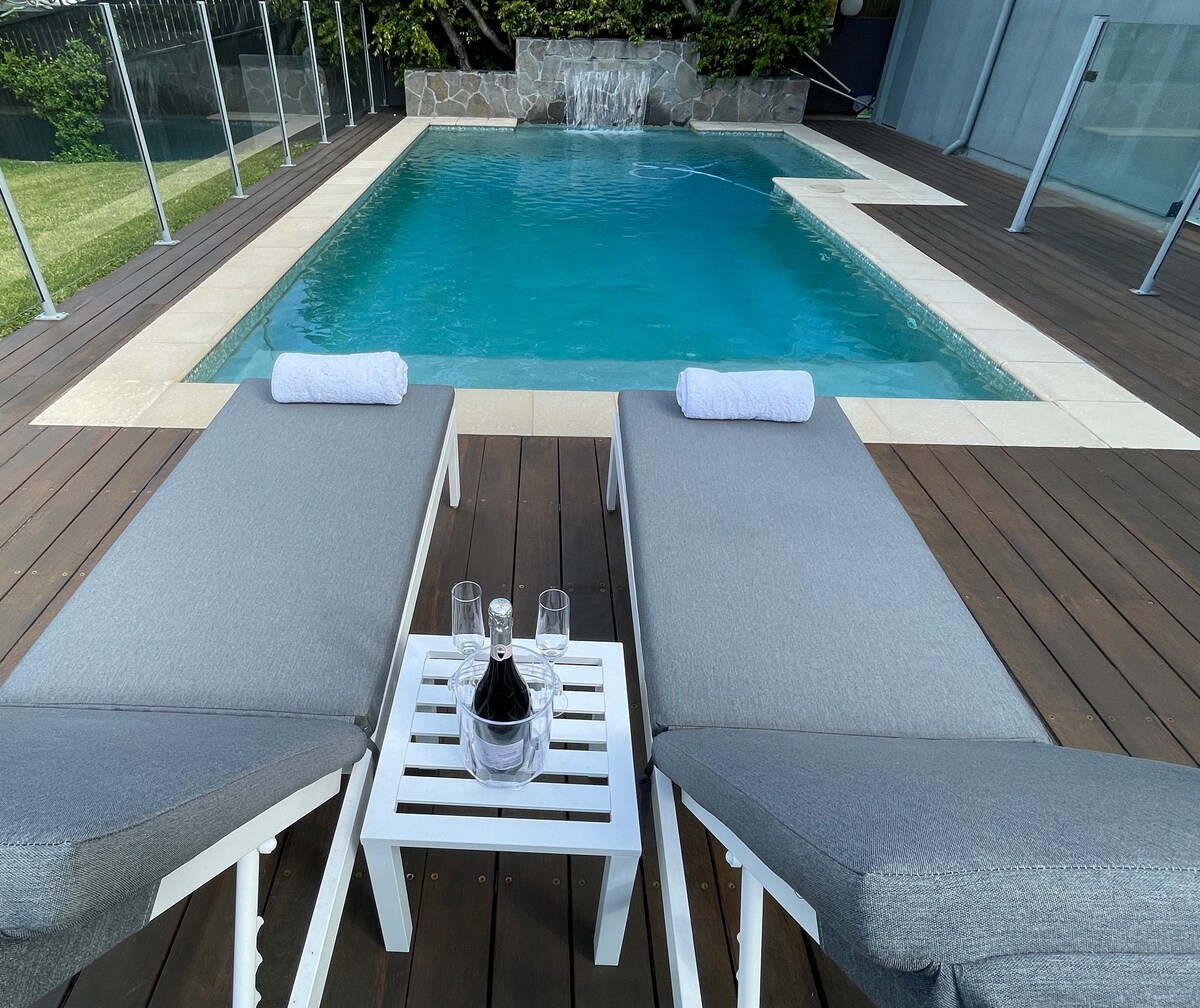 Two sun loungers with grey cushions are positioned beside a sparkling saltwater pool, featuring a serene waterfall. A small white table sits between them, holding a bottle of champagne and two glasses. The pool area is framed by landscaped greenery.