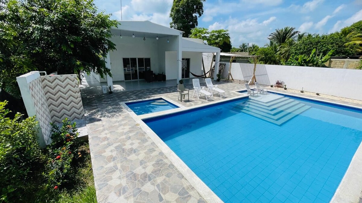 A spacious outdoor area is highlighted by a large swimming pool with shimmering blue water. Surrounding the pool are several lounge chairs on a patterned stone deck. A nearby pavilion offers shade and includes a hammock. Lush greenery is present in the background.