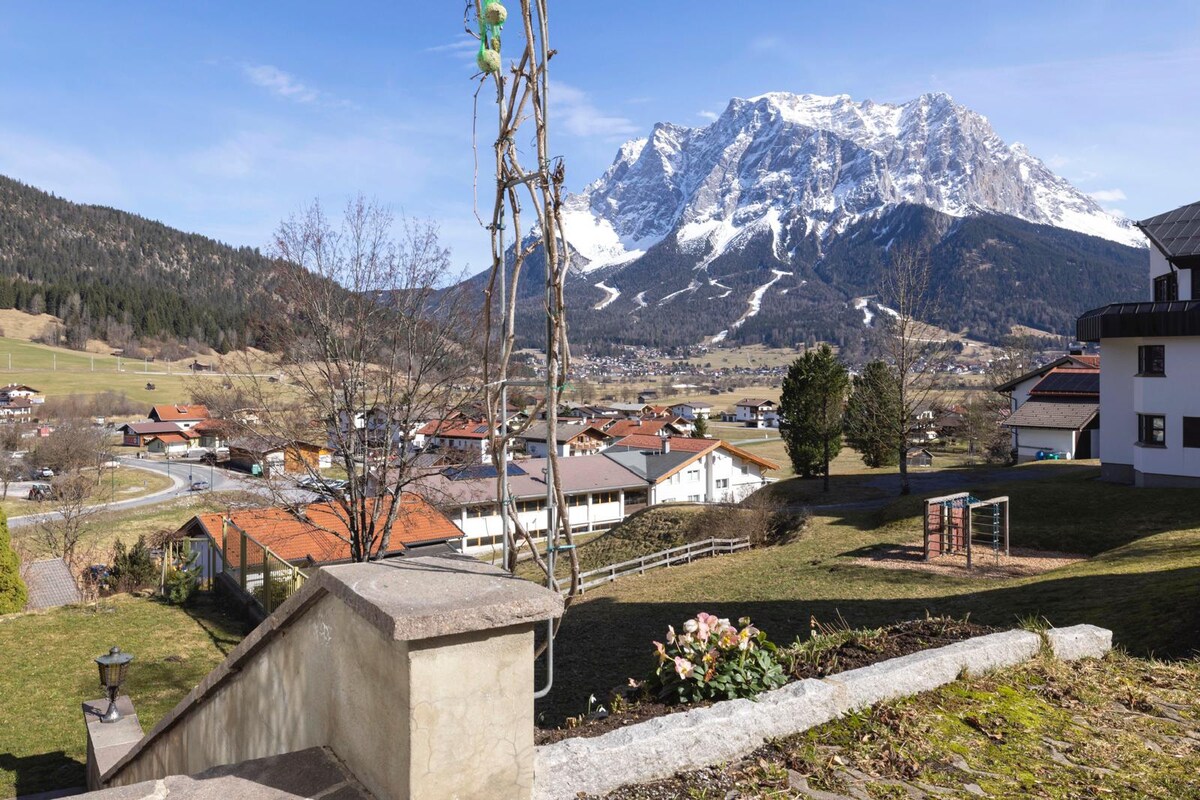 A scenic view of the surrounding landscape is displayed, featuring the majestic Zugspitze mountain in the background. The image showcases a well-maintained garden with budding flowers and a pathway leading up to steps. Nearby homes and greenery are visible, enhancing the tranquil atmosphere.