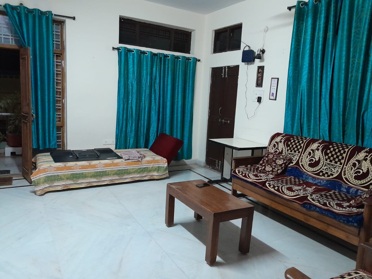 A cozy living area features a brown wooden coffee table at the center, surrounded by a patterned sofa and a foldable bed with colorful bedding. Windows are framed by bright teal curtains, while the floor is covered in light-colored tiles.