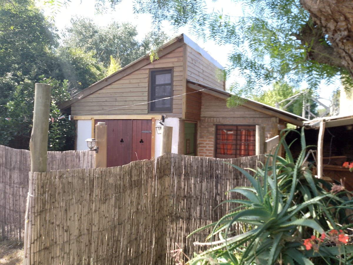 A two-story cabin features wooden exterior accents, nestled among greenery. A small fence made of bamboo provides a natural boundary, while large windows allow for natural light. The roof has a gentle slope, indicating a welcoming outdoor space.