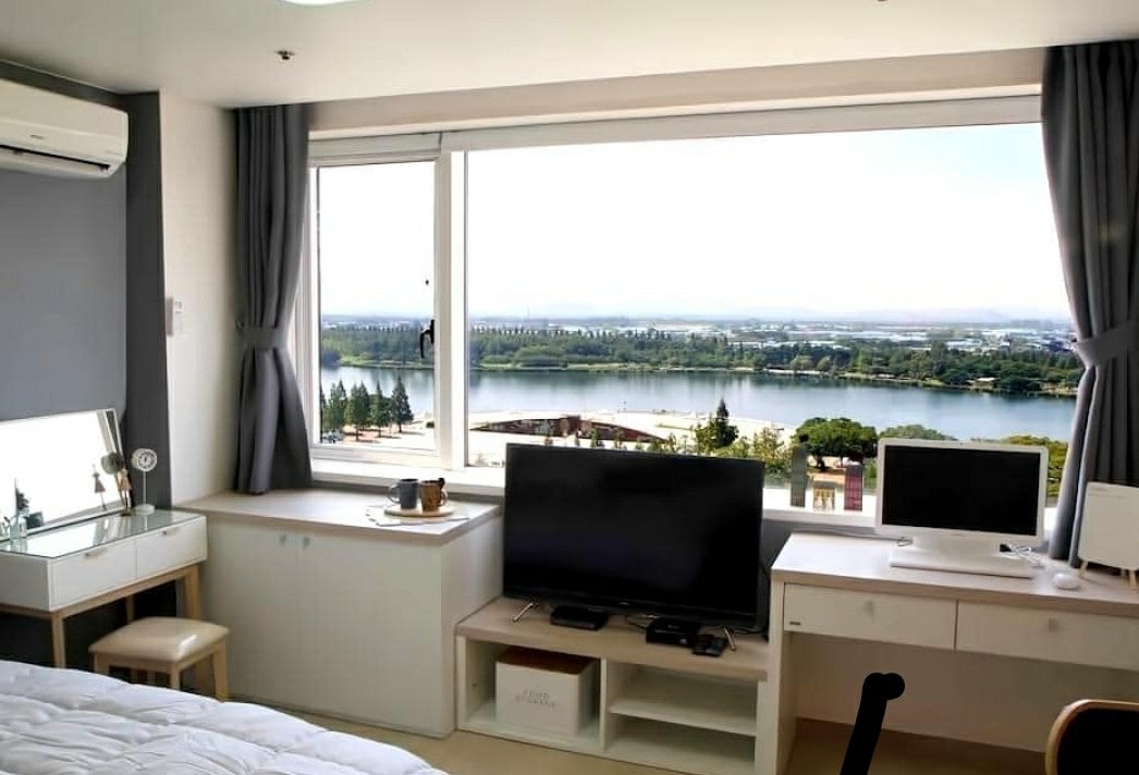A bright room features large windows offering views of a serene body of water and greenery. A spacious bed is complemented by a mirrored vanity and a modern television setup on a white console beneath the window. Elegant curtains frame the scene.