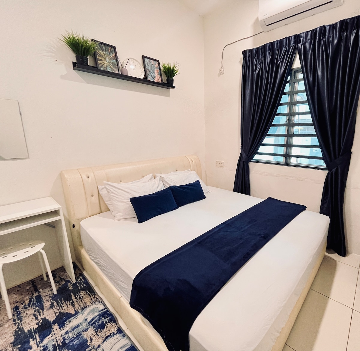 A spacious bed is positioned against a light-colored wall, featuring dark blue bedding. A streamlined window with horizontal bars allows natural light to enter. A small desk with a chair is visible nearby, complemented by decorative items on a shelf above the bed.