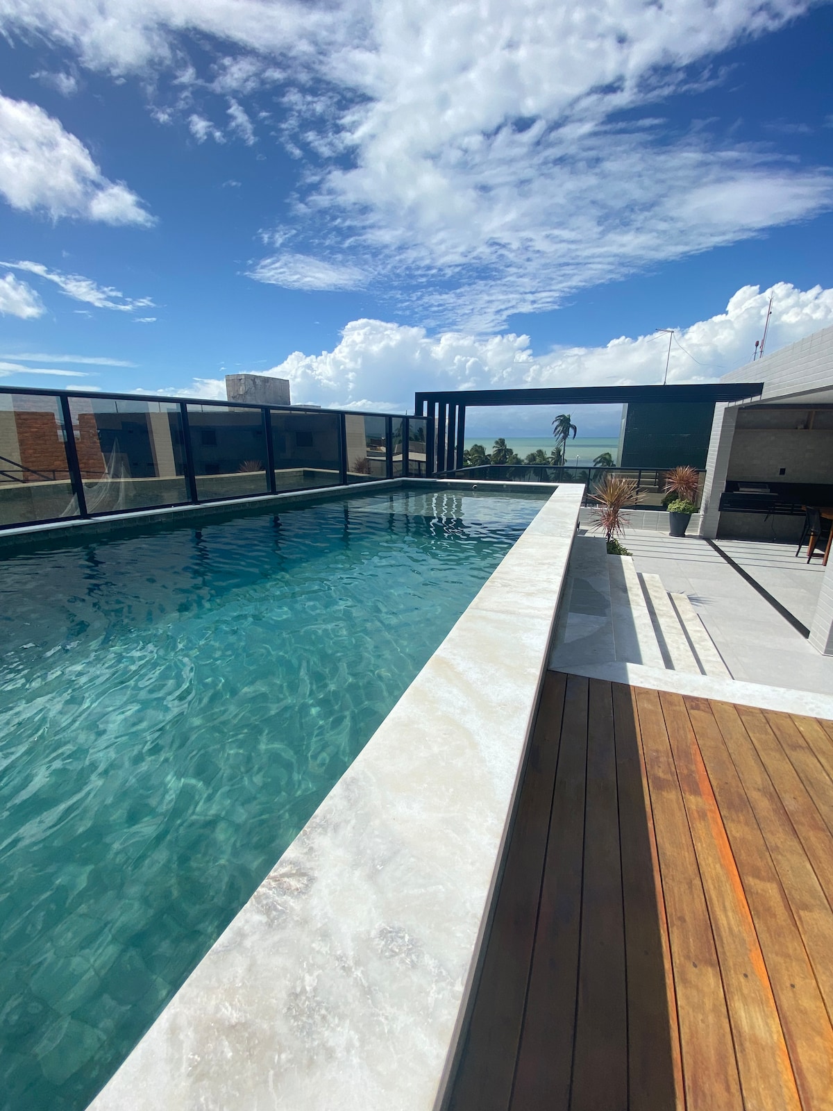 A rooftop pool area features a shimmering body of water with a clear blue sky above. Surrounding elements include a wooden deck and glass railings, providing unobstructed views of the landscape. Sunlight reflects off the water's surface, creating a tranquil environment.