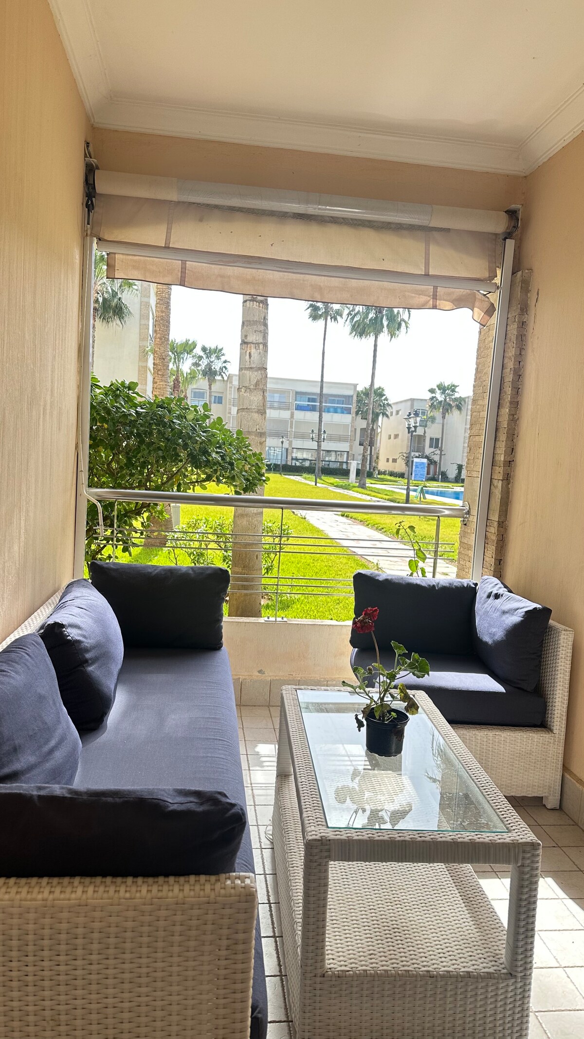 A charming terrace is visible, featuring a comfortable seating arrangement with dark cushions on a wicker sofa set. A glass coffee table centers the space, adorned with a potted plant. Lush greenery and palm trees create a serene backdrop, enhancing the outdoor ambiance.