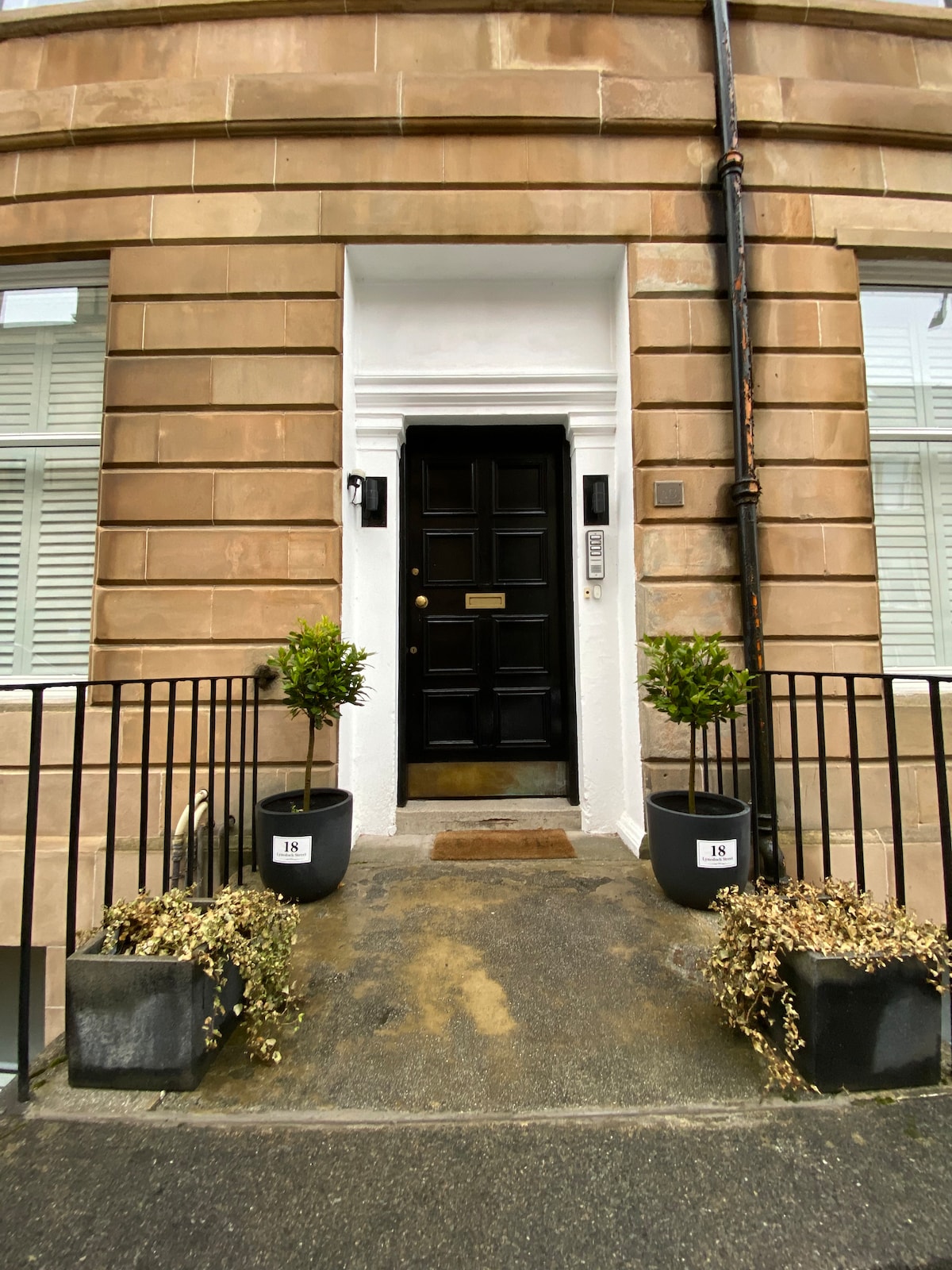 The entrance of the flat features a dark wooden door framed by two potted plants. The path is bordered by a low black railing, and the address number is displayed on the wall beside the door, providing a welcoming entry point.