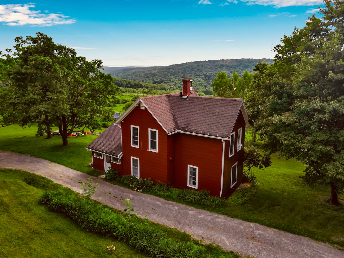 A charming country house is set against a backdrop of rolling hills and verdant trees. The red exterior contrasts with the lush green landscape, and a winding gravel driveway leads to the entrance. Surrounding gardens exhibit well-maintained plants, enhancing the serene atmosphere.