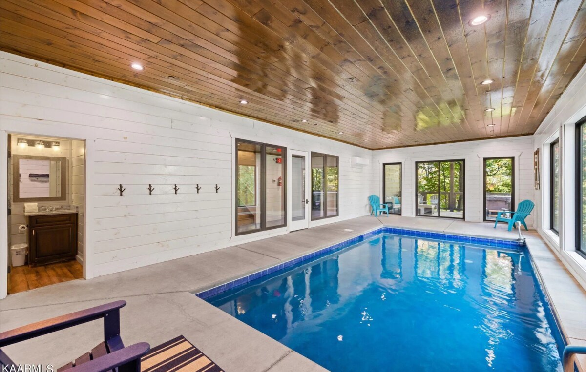An indoor heated pool is surrounded by large windows creating a bright and airy space. The wooden ceiling adds warmth, while two blue chairs provide seating. An area for changing with hooks is visible, enhancing convenience.