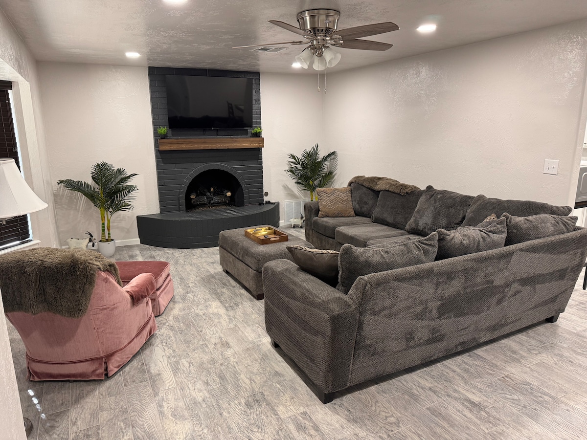 A spacious living room is furnished with a large sectional sofa and a comfortable accent chair. A coffee table is positioned in front of the sofa. A decorative fireplace with a wooden mantel serves as a focal point, while a ceiling fan provides ventilation.