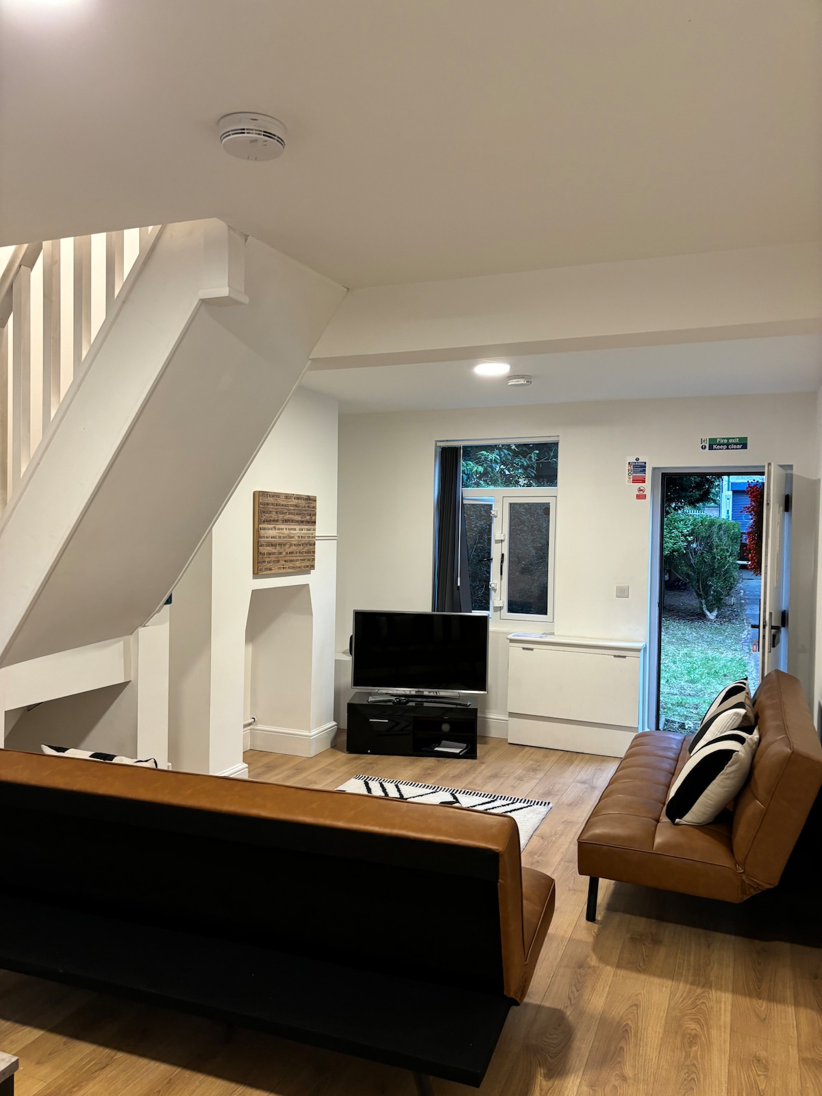 An inviting living space features a leather sofa and a modern television, set against light wooden flooring. Natural light floods in through the large window, which offers a view of the green exterior. A staircase elegantly leads to the upper level.