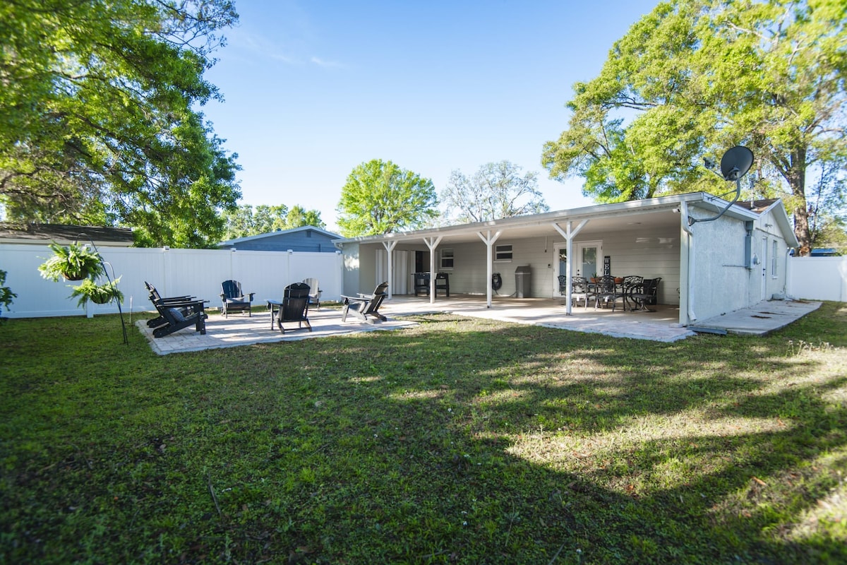 A spacious backyard is depicted, featuring a covered patio area with seating arranged for relaxation. Several chairs are positioned around a stone-paved space, and a dining table with chairs is visible. The yard is surrounded by lush greenery and a privacy fence.