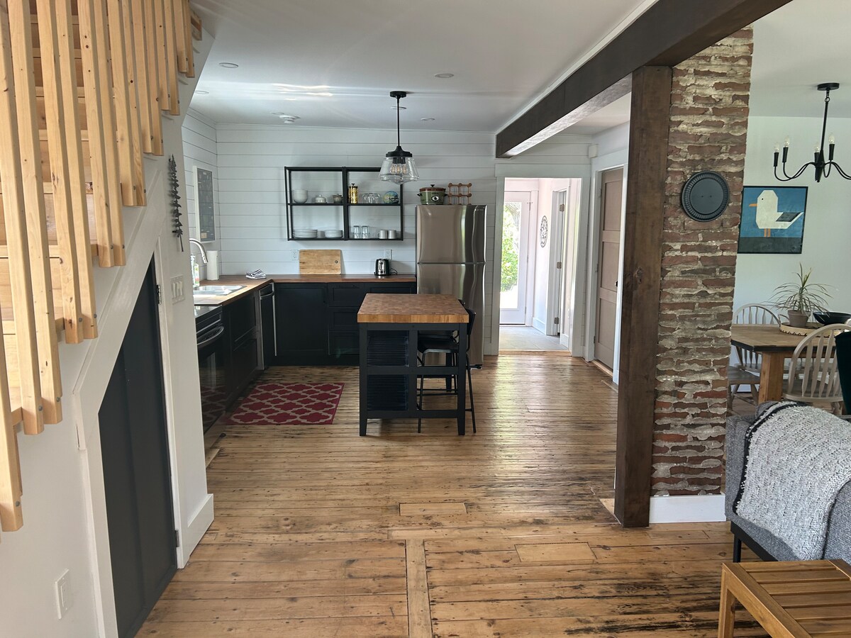 A modern kitchen area features dark cabinetry and stainless steel appliances. A central island provides additional workspace and seating. Exposed wooden beams and original hardwood flooring enhance the rustic charm, while natural light flows in from nearby windows.