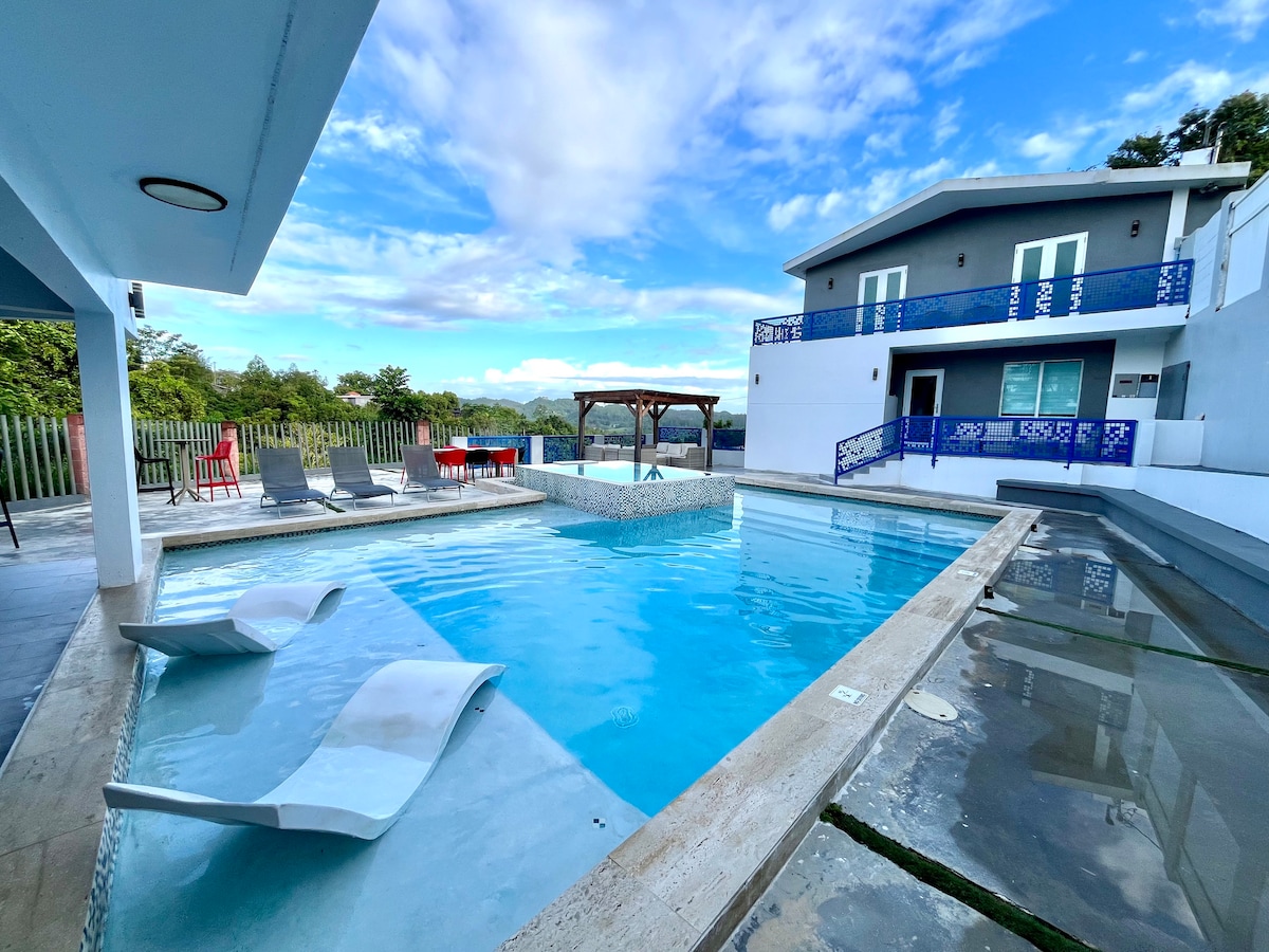 A large heated pool is surrounded by a spacious sun deck, featuring lounge chairs and a shaded pergola. The water reflects the sky, while nearby, a modern building offers multiple levels with blue accents and outdoor seating for relaxation.