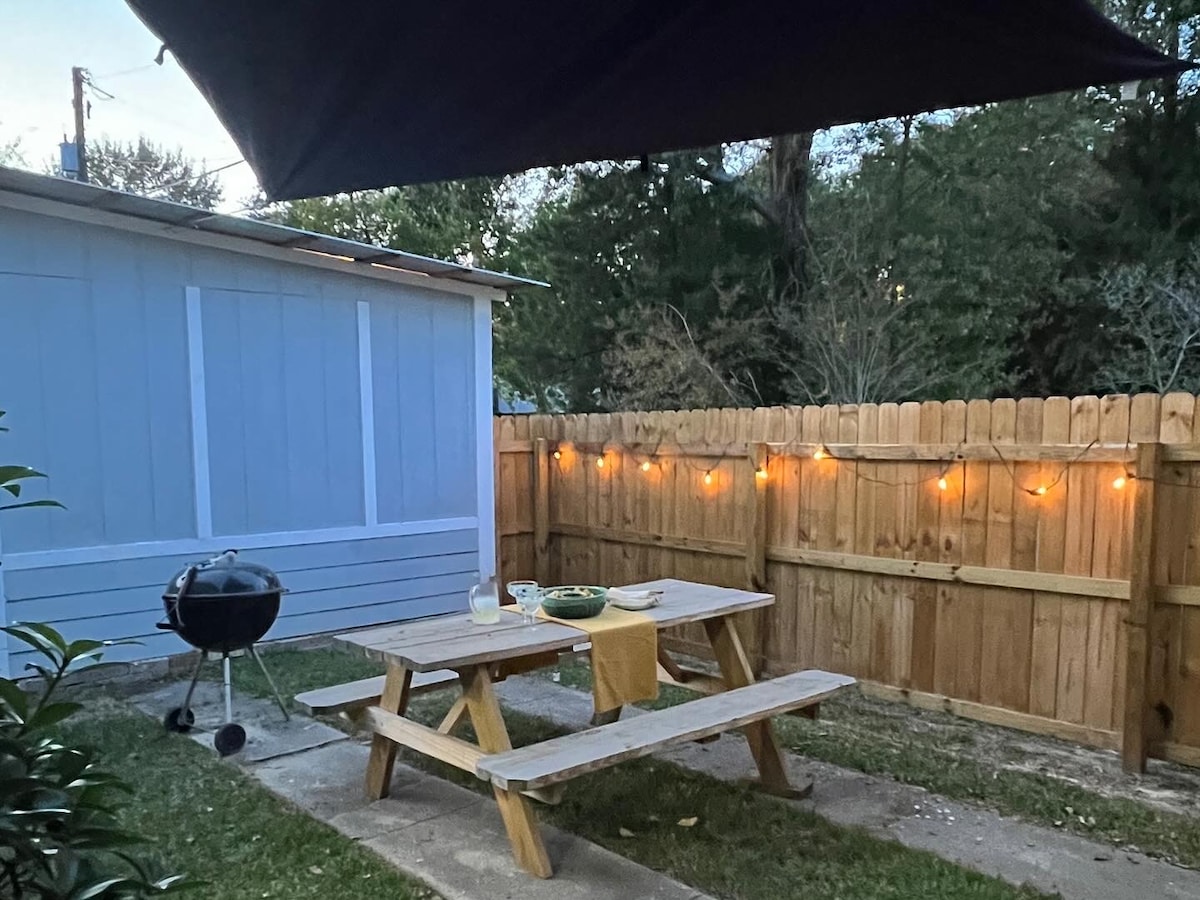 An outdoor seating area is shown, featuring a wooden picnic table set under a shaded awning. Soft lights are strung along the wooden fence, creating a cozy ambiance. A grill is positioned nearby, surrounded by a well-kept garden area.