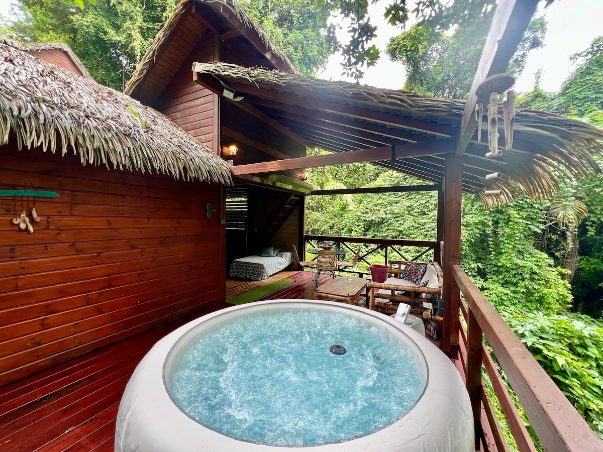 An outdoor terrace features a circular spa surrounded by lush greenery. The wooden structure showcases a rustic charm with a thatched roof, while a glimpse of a cozy sleeping area is visible in the background. Natural light filters through the surrounding foliage.