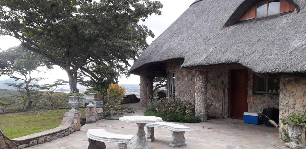 A stone patio features a circular table surrounded by two curved benches, providing seating under a thatched roof. Lush greenery and large trees are visible in the background, enhancing the outdoor setting. A distant view of the landscape can be seen beyond the patio.