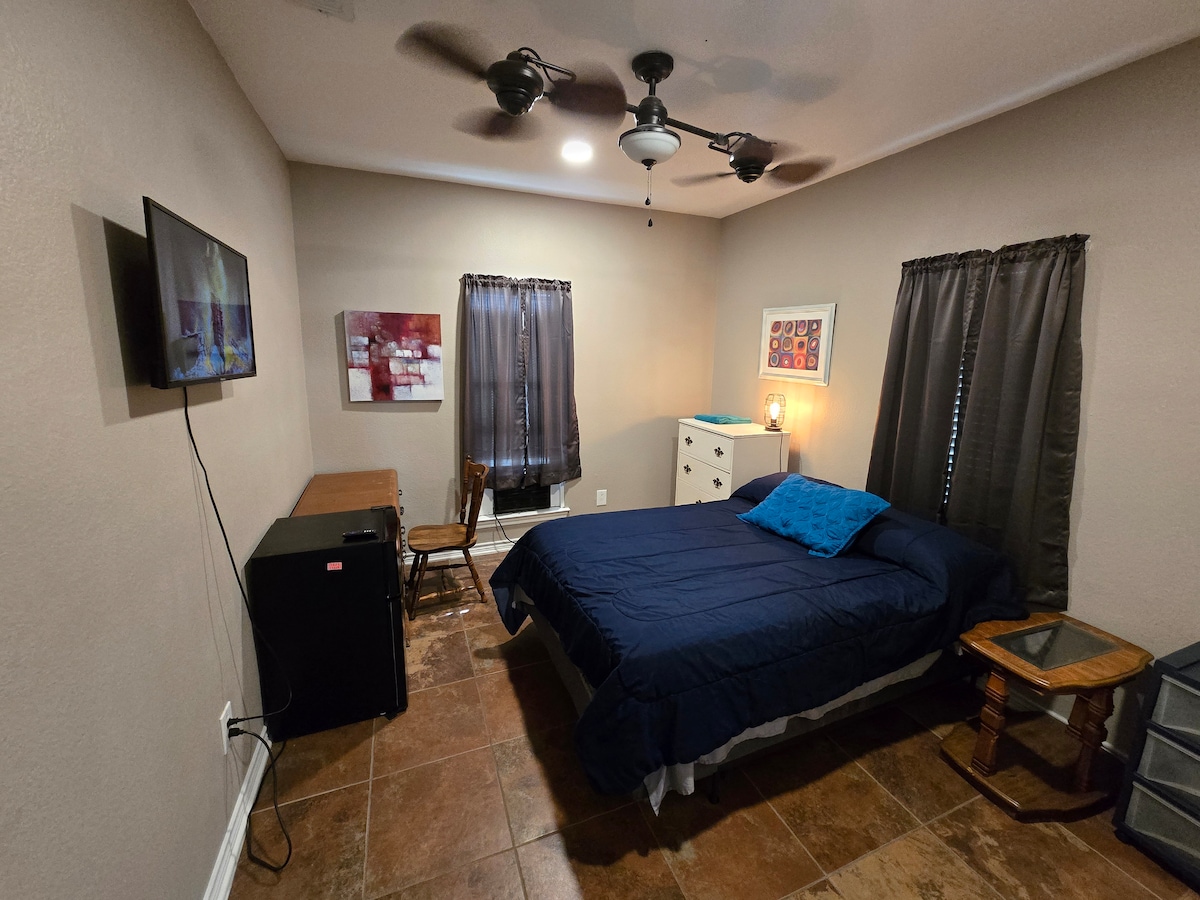 A private room features a cozy double bed with dark bedding and a blue accent pillow. A television is mounted on the wall, complemented by a mini fridge and a desk with a wooden chair. Two windows with dark curtains allow for controlled lighting.