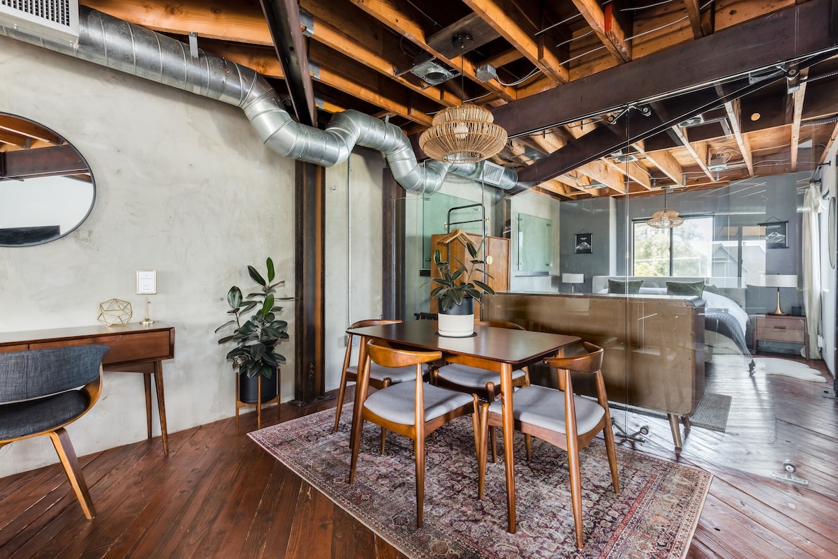 A dining area features a wooden table surrounded by four chairs, complemented by a decorative rug. Exposed beams and ductwork enhance the industrial style. Natural light fills the space through large glass walls, revealing the adjacent kitchen and bedroom area.