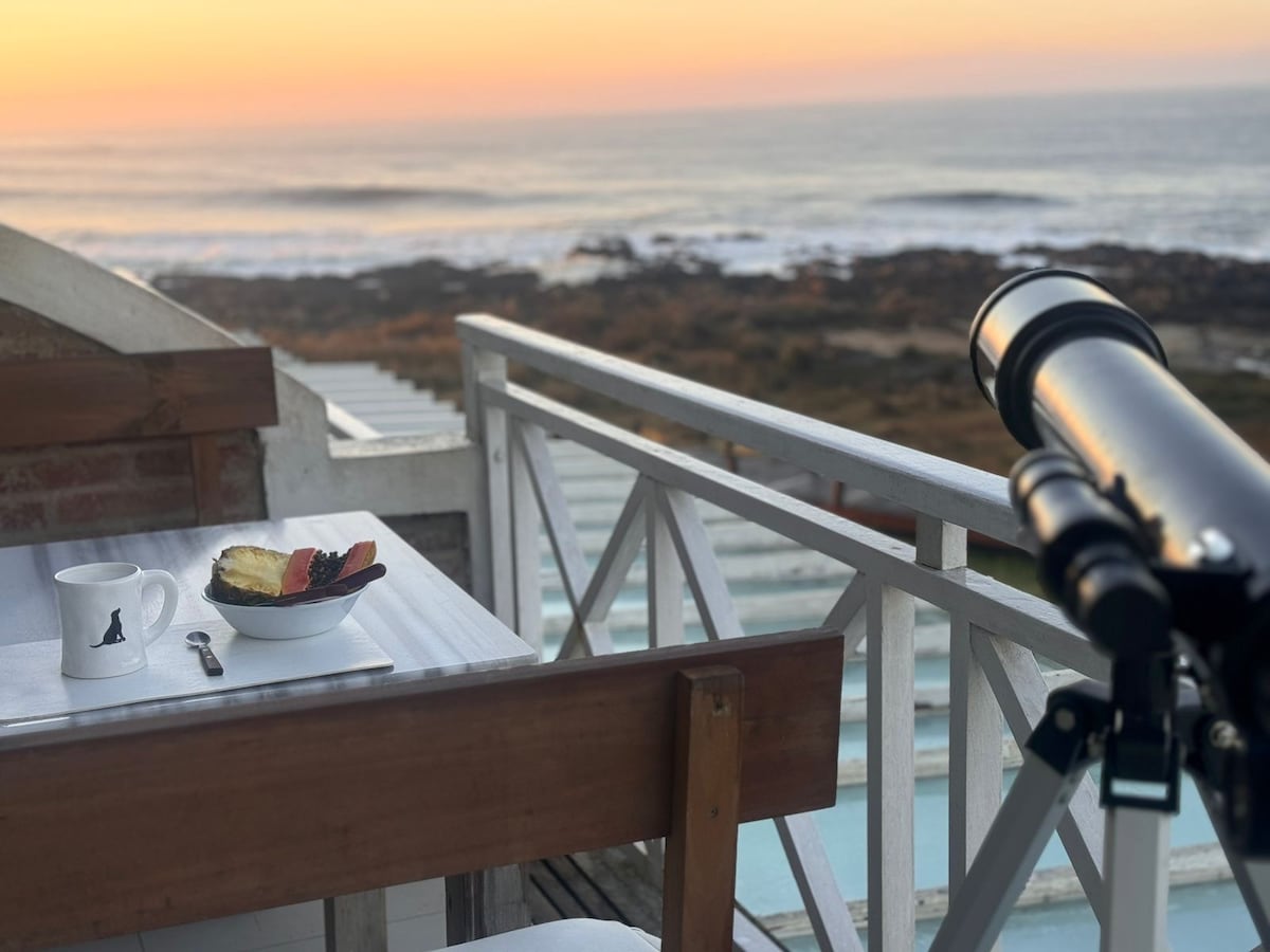 A terrace table is set with a bowl of fruit and a coffee cup, overlooking the ocean. A telescope stands nearby, offering a view of the serene coastline at sunset, where the sky is painted with soft hues of orange and pink.