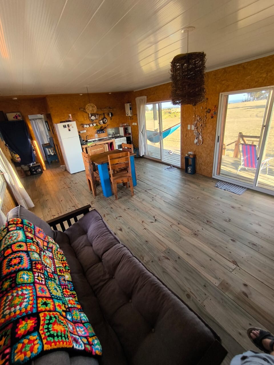 An inviting living area is showcased with wooden floors and a chunk of natural light streaming through large glass doors. A brown sofa is draped with a colorful crocheted blanket, while a dining table with chairs is positioned centrally, creating a cozy atmosphere.