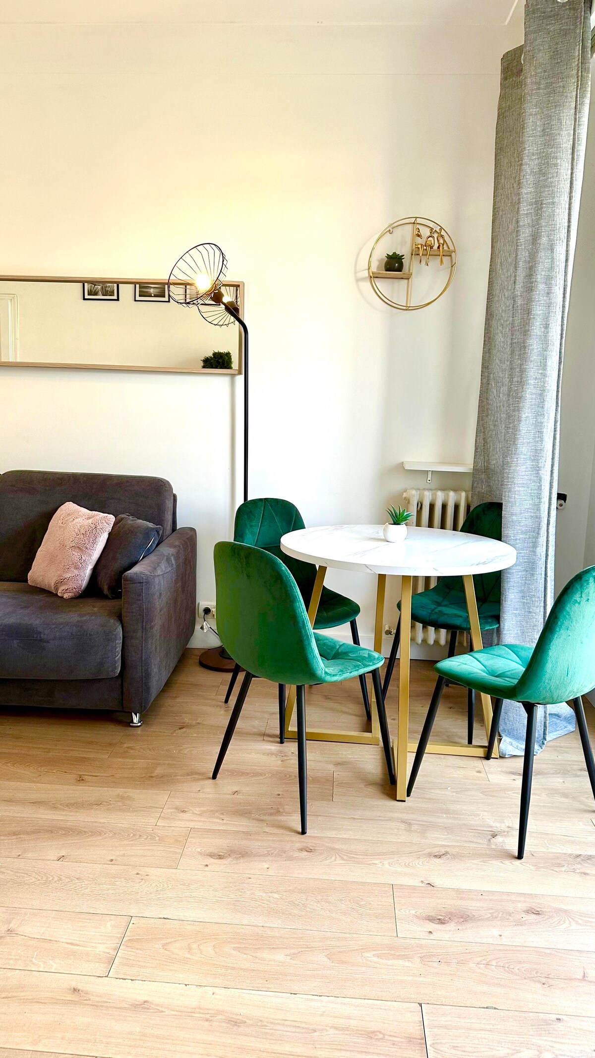 A cozy dining area is highlighted by a round white table surrounded by four green velvet chairs. A gray sofa sits nearby, while light wood flooring enhances the space. A large mirror reflects the area, and a decorative wall feature adds a touch of elegance.