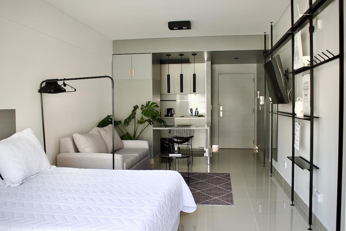 A modern studio area features a spacious bed with crisp white linens and a cozy grey sofa. A small kitchenette is visible in the background, equipped with sleek cabinetry and essential appliances. Natural light illuminates the polished floor, complemented by a simple area rug and decorative plant.