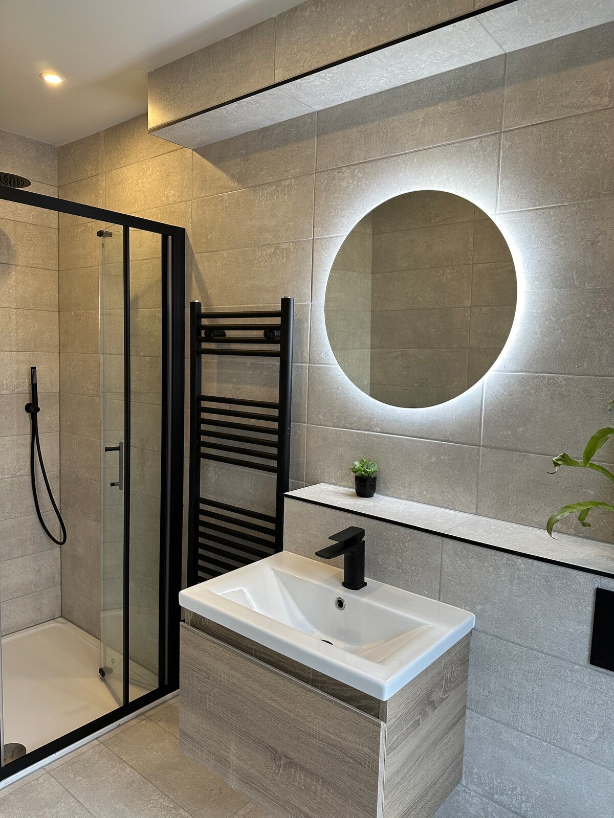 A modern bathroom features neutral tile walls and a sleek, minimalistic design. A round mirror with backlighting hangs above a compact sink. A tall towel radiator, a glass shower enclosure with black fixtures, and a small plant add subtle touches of detail.