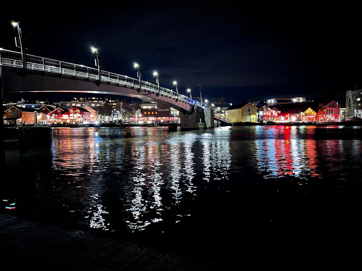 A well-lit bridge spans across the water, reflecting colorful lights from nearby buildings. The scene captures a serene nighttime ambiance, with illuminated structures contributing to a lively waterfront atmosphere. The still water mirrors the vibrant hues, enhancing the visual appeal of the surroundings.