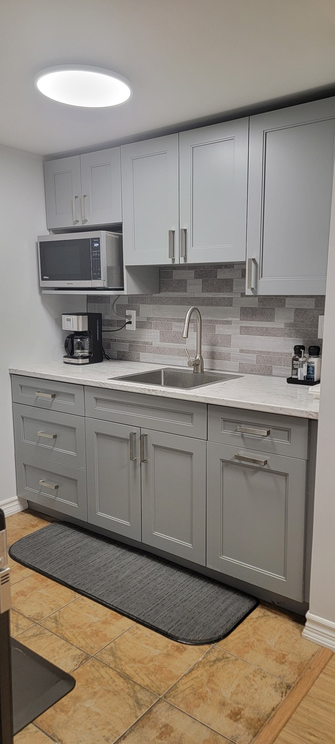 A modern kitchen area features light grey cabinetry with ample storage space. A white countertop surrounds a stainless steel sink, while a microwave and coffee maker are positioned on the countertops. A subtle grey mat is placed on the floor, enhancing the functionality of the space.