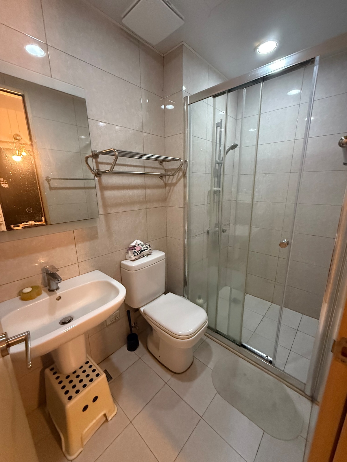 A compact bathroom is equipped with a glass shower enclosure, a modern toilet, and a small sink. Wall-mounted shelves provide storage space. A illuminated mirror enhances visibility. A step stool is positioned beside the sink, offering convenience.