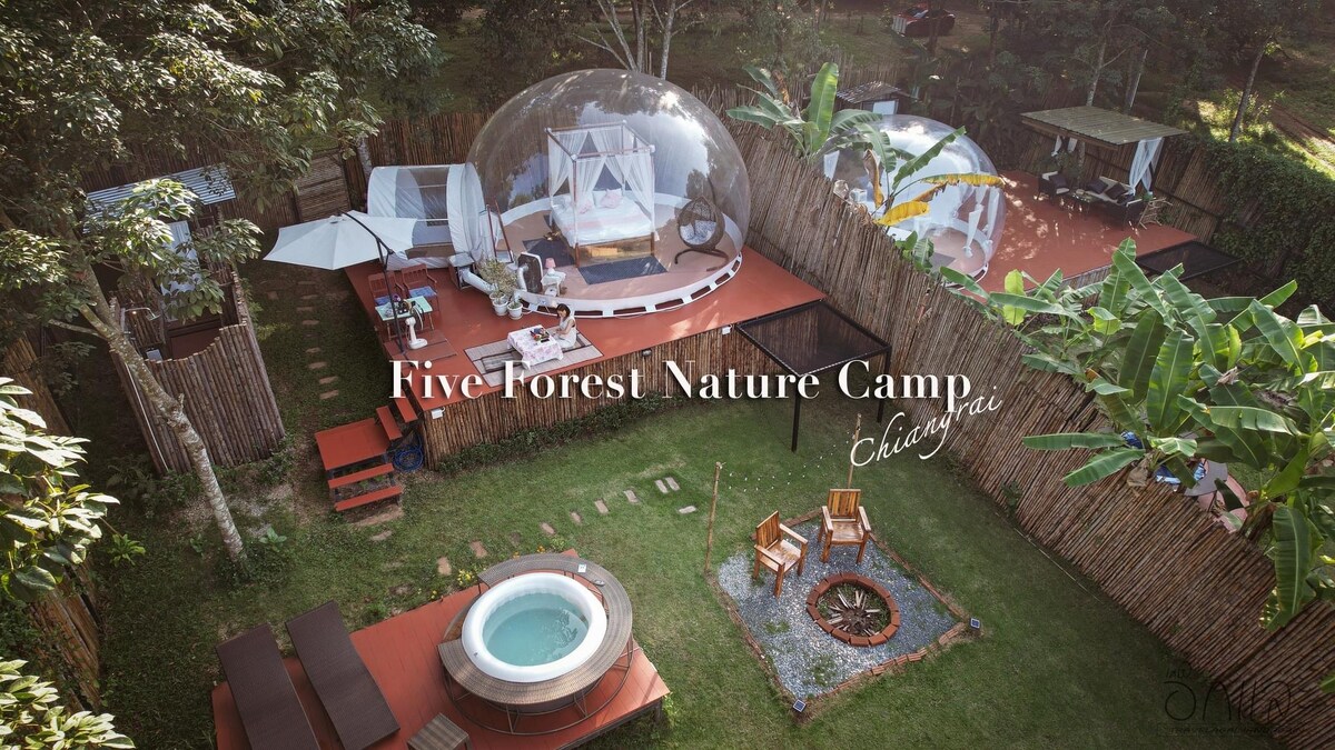 A nature camp setup is visible, featuring clear bubble accommodations surrounded by lush greenery. Outdoor seating areas include a fire pit and a hot tub, with natural elements integrating into the design. Wooden decking enhances the spacious outdoor environment.
