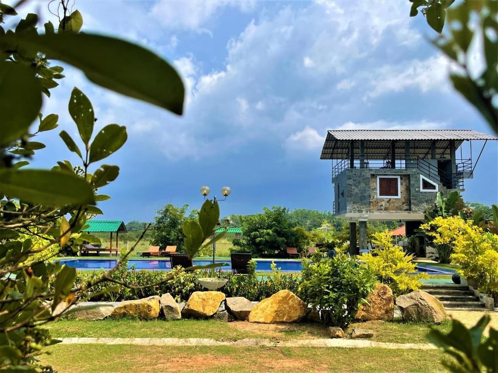 A serene view of a modern villa emerges among lush greenery and boulders. The two-story structure features a large terrace and is positioned near a sparkling pool, surrounded by lounge chairs and vibrant landscaping under a partly cloudy sky.