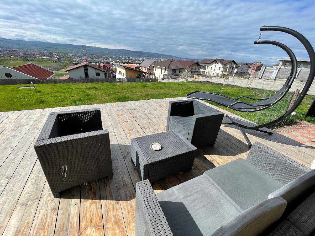 An outdoor seating area features modern wicker furniture arranged around a small table. A gently sloping landscape with homes is visible in the background, and a hammock stands nestled among the grass, with clouds drifting in a blue sky.