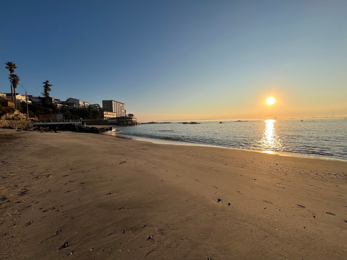 The image captures a serene beach at sunset, with soft sand in the foreground leading to calm water. A soft orange glow is reflected on the water's surface as the sun descends on the horizon, with a distant building and trees adding to the scenic backdrop.