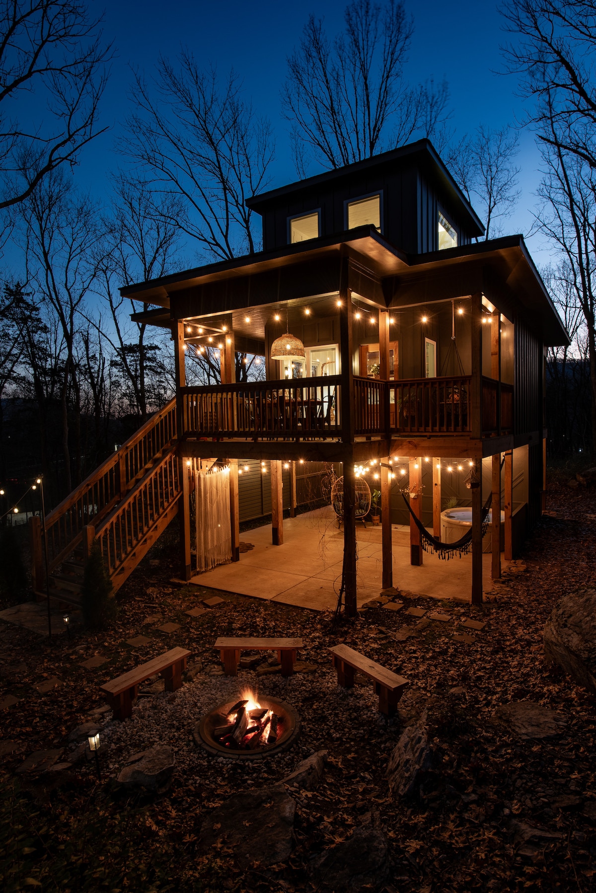 The treehouse retreat is softly illuminated by string lights, creating a warm atmosphere during twilight. A fire pit surrounded by natural stone seating is positioned in front, with a winding staircase leading up to the spacious deck adorned with inviting seating options.