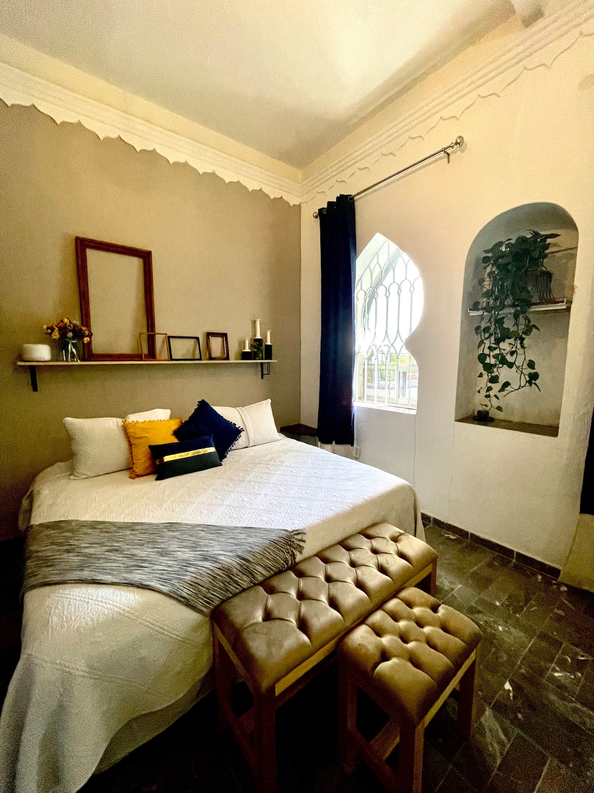 A spacious bedroom features a comfortable bed dressed in neutral linens, flanked by decorative pillows. Two golden benches sit at the foot of the bed. Natural light filters through an arched window, highlighting the warm wall tones and minimalist decor.